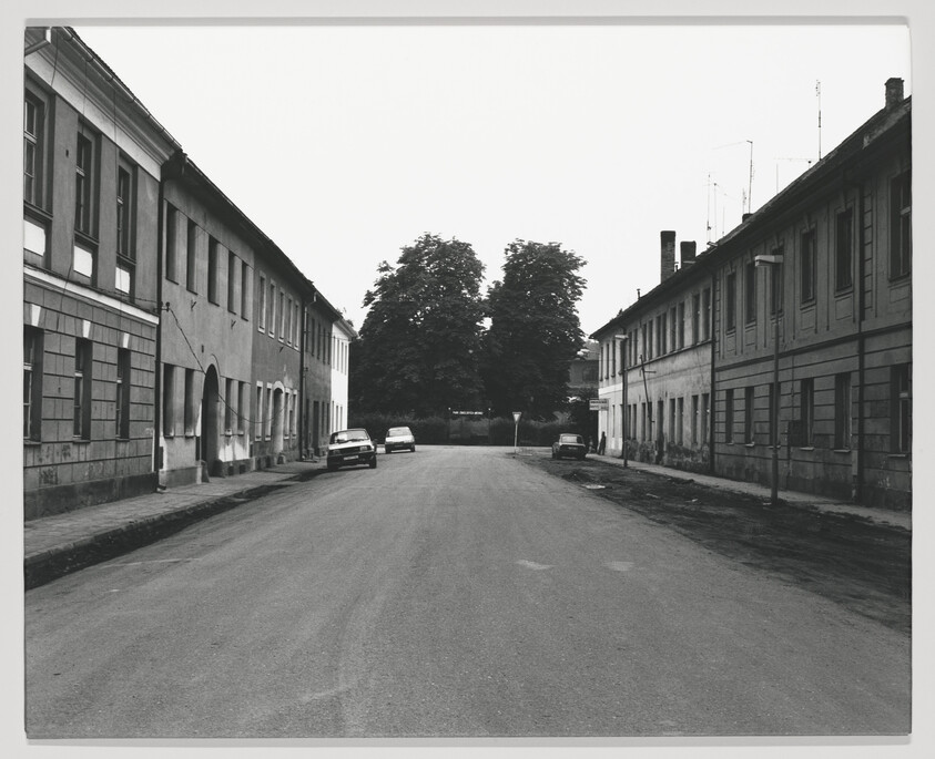 A black and white photograph of a quiet street lined with two-story buildings, some with boarded windows. A few cars are parked along the sides of the road, and there are trees visible at the far end of the street.
