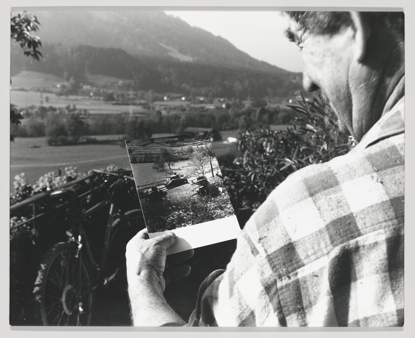 A black and white photo showing a person from behind, looking at an old photograph they are holding in their hands. The photograph within the photo depicts a rural scene with trees and a small body of water. In the background, a mountainous landscape is slightly out of focus, suggesting the person is comparing the current view with the one in the photograph. The person is wearing a plaid shirt, and the edge of a bicycle is visible in the lower left corner of the image.