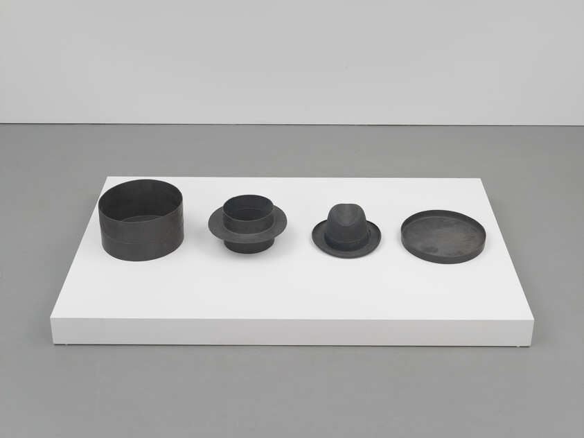 Four dark circular metal forms resembling hat components sit in a row on a white pedestal.