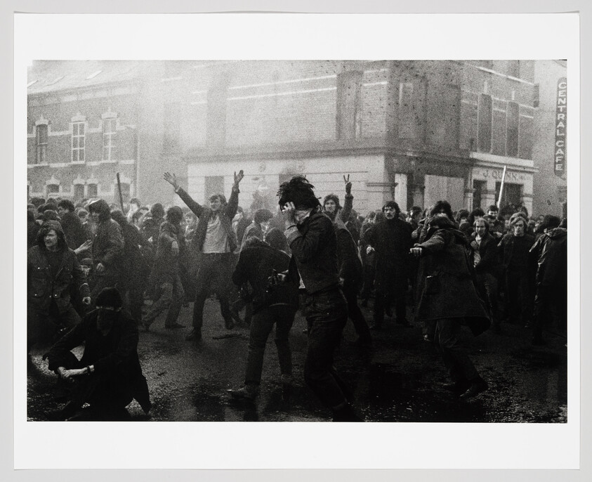 A large crowd of protesters clashes in a wet street, some people running and raising hands.