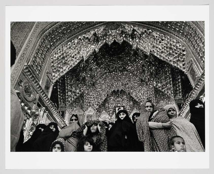 A black and white photograph capturing a group of people, predominantly women wearing hijabs, standing in front of an ornately decorated archway with intricate mosaic patterns. The expressions on their faces range from solemn to curious, as they look in various directions. The mosaic creates a shimmering backdrop that contrasts with the subjects' dark attire.