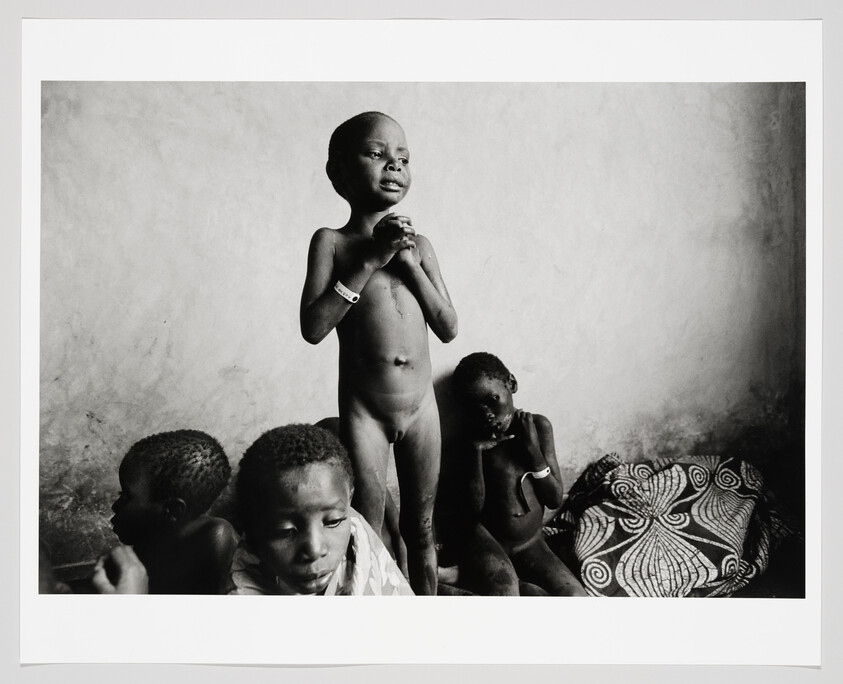 A group of severely malnourished children in a bare room, one child standing and clasping hands.