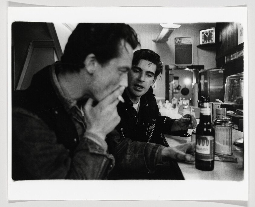 Two young men sit at a diner counter, one smoking and the other holding a beer.