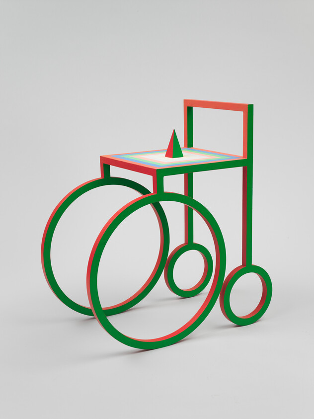 A minimalist red-and-green geometric sculpture resembling a chair with large circular wheels and a cone.