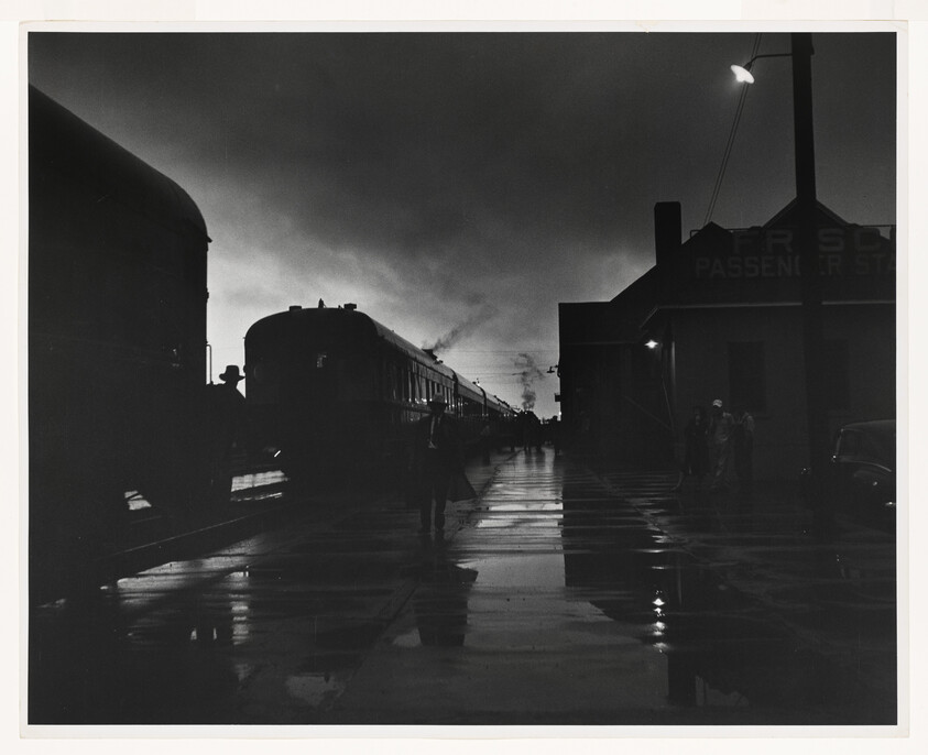 Silhouetted passengers walk along a wet platform beside a steam train at dusk.