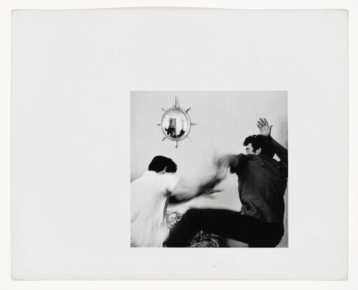 Two men struggle physically in a room, one raising an arm while the other swings at him.