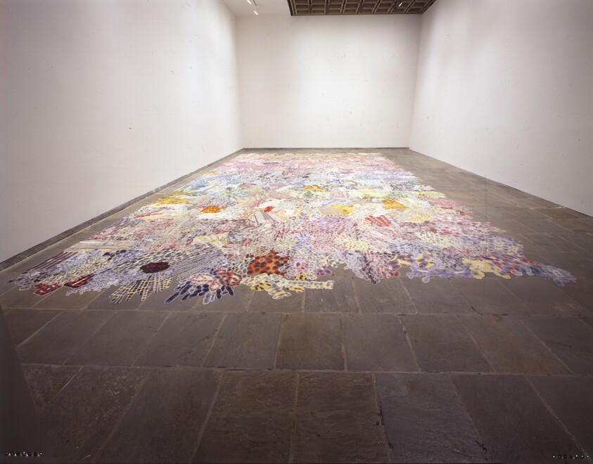 A large colorful patchwork-like floor installation spreads across a gallery room.