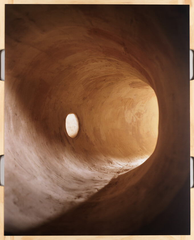 Interior view of a curved wooden tunnel structure with warm lighting, showcasing the smooth texture of the wood and the circular patterns created by the construction. The tunnel narrows into a bright, oval-shaped opening at the far end.