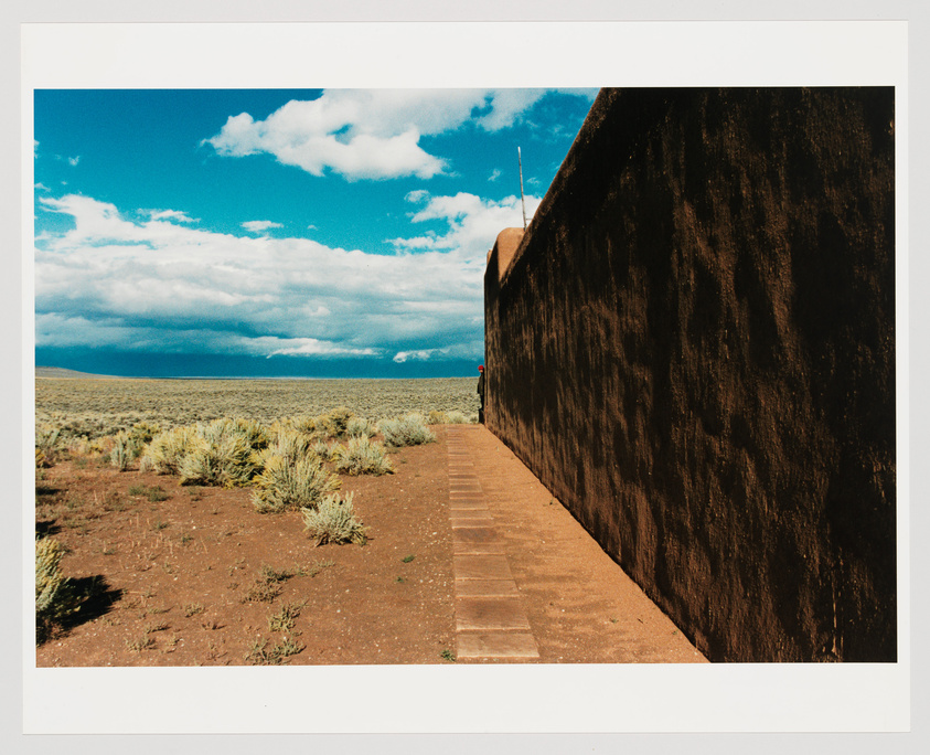 A long, dark brown adobe wall stretches from the foreground to the middle of the image on the right side, leading to a building with a rounded corner and a small red object at the base. A brick pathway runs alongside the wall. The landscape opens to a vast, flat desert with sparse shrubbery under a wide sky filled with fluffy white clouds.