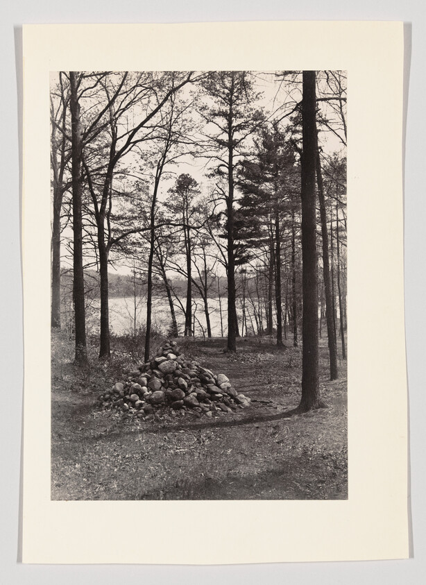 A large pile of stones sits in a quiet forest clearing beside a lake.