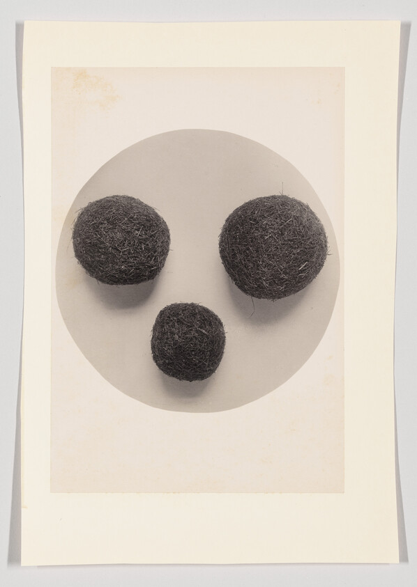 Three dark felt balls arranged like a face on a circular gray background mounted on paper.