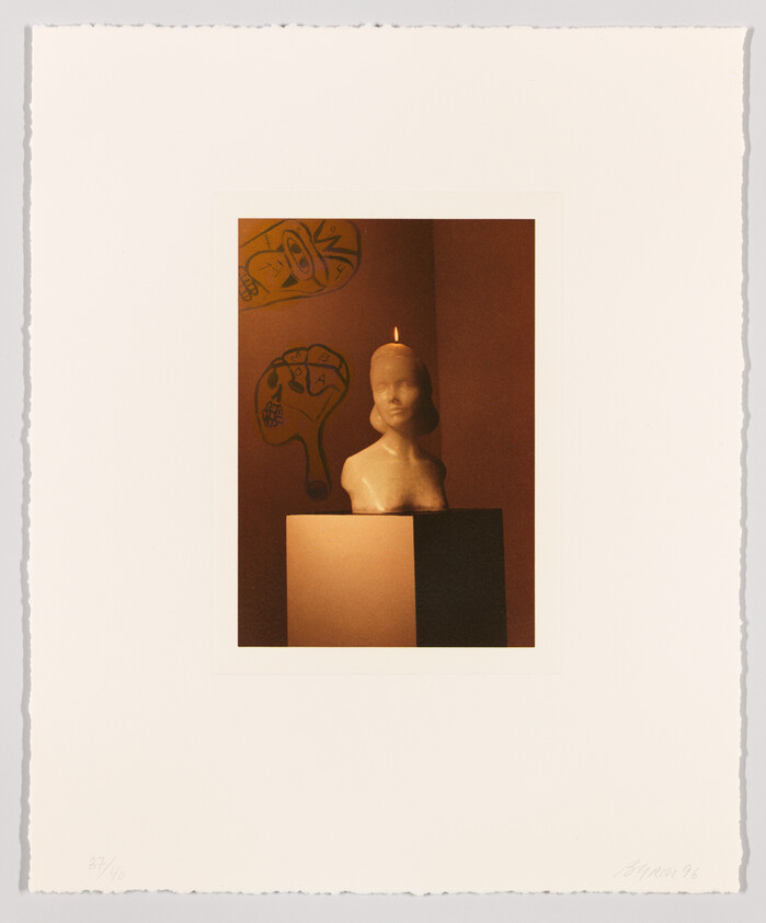 A wax bust with a small flame on its head sits on a pedestal against a wall with drawings.