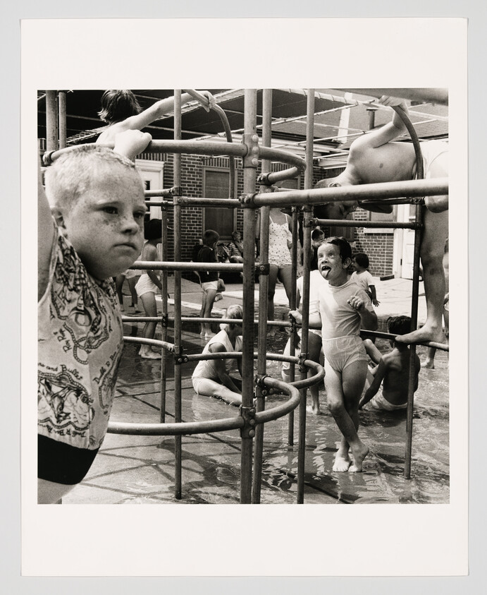 Several children play and climb on a metal jungle gym in shallow water at a pool.