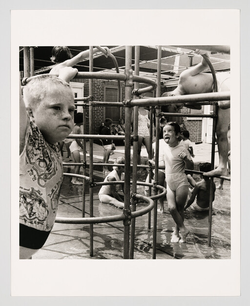 Children play on a metal jungle gym in shallow water while one boy sticks out his tongue.
