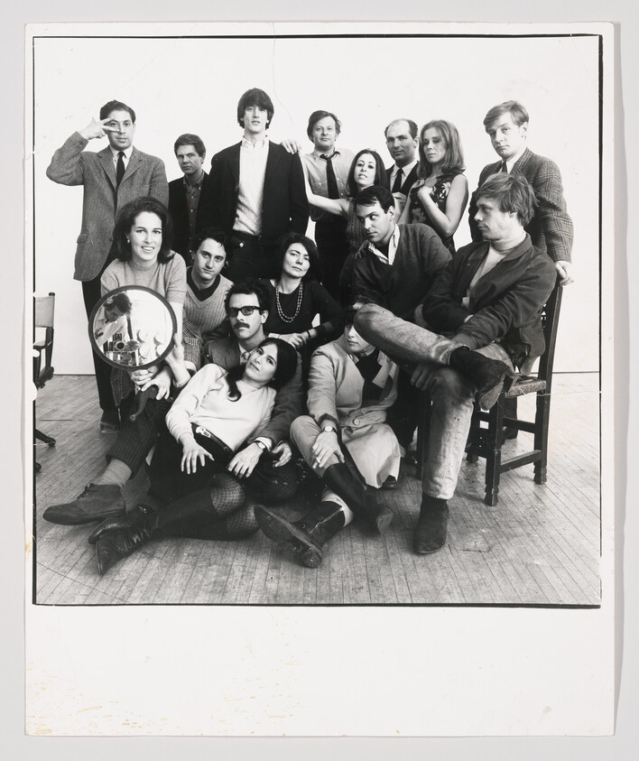 A large group of young adults pose together in a black-and-white studio photo, one holding a round mirror.