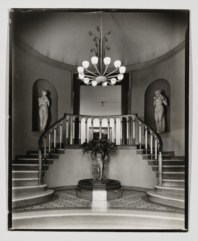 Grand curved staircase with central sculptural fountain, flanked by two niche statues and chandelier.