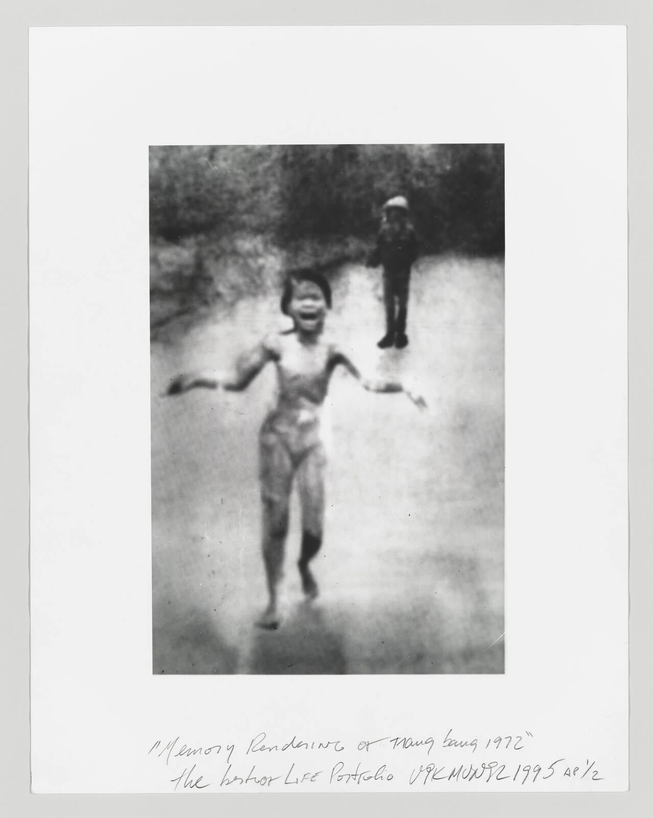 A young naked child runs down a road with arms outstretched while a soldier stands behind.