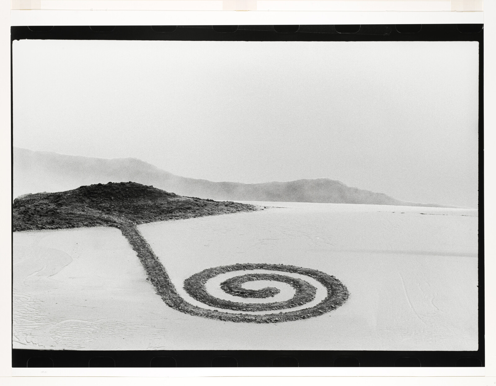 A long stone spiral path winds from a low mound across a flat salt plain toward distant hills.