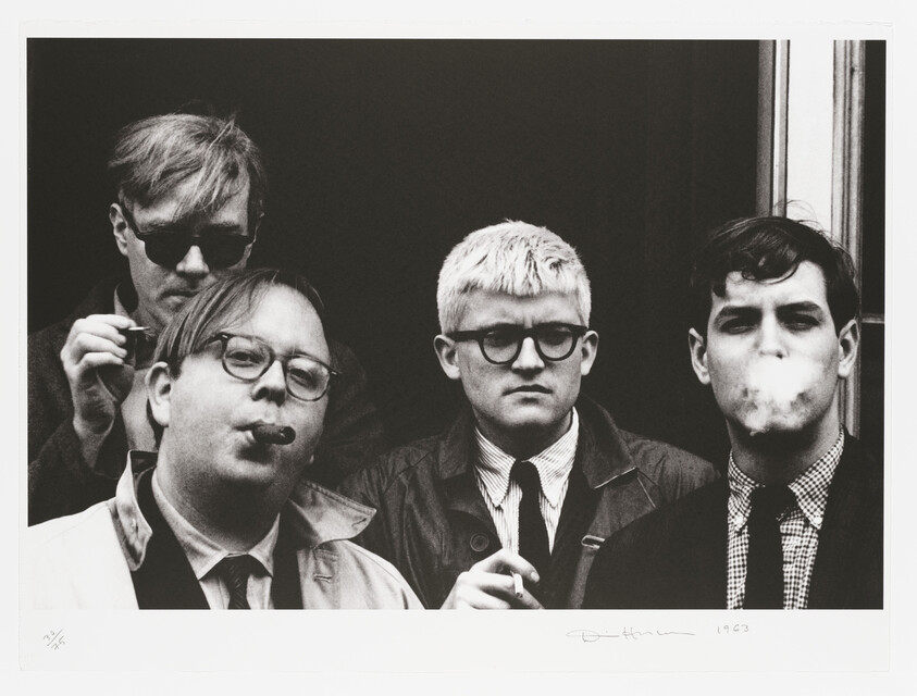 A black and white photograph of four men posing with serious expressions. The man on the far left wears sunglasses and is lighting a cigarette, the second from the left holds a pipe in his mouth, the third man is wearing a checked shirt and a jacket, and the man on the far right is exhaling smoke from a cigarette. They all have short haircuts and are standing in front of a dark background. There is a signature and the year "1963" written in the bottom right corner of the image.