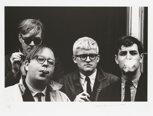 Four young men in jackets and glasses stand together, two smoking while one exhales a cloud of smoke.