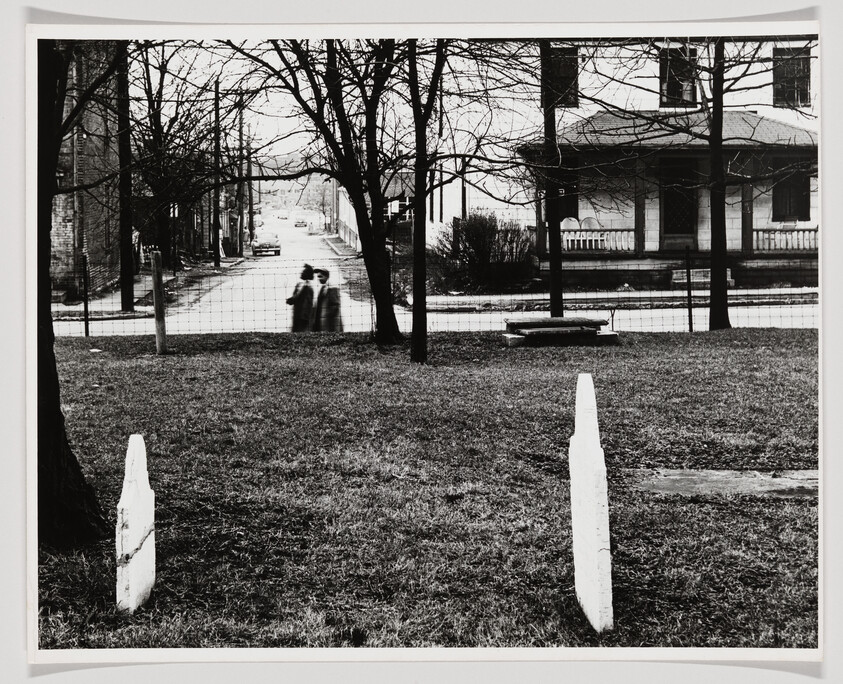 Two white grave markers in a yard with two blurred people walking along the street behind.
