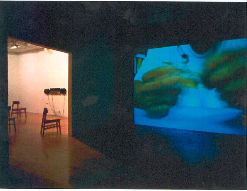 A dark room with a large video projection of hands, next to a lit room with empty chairs.