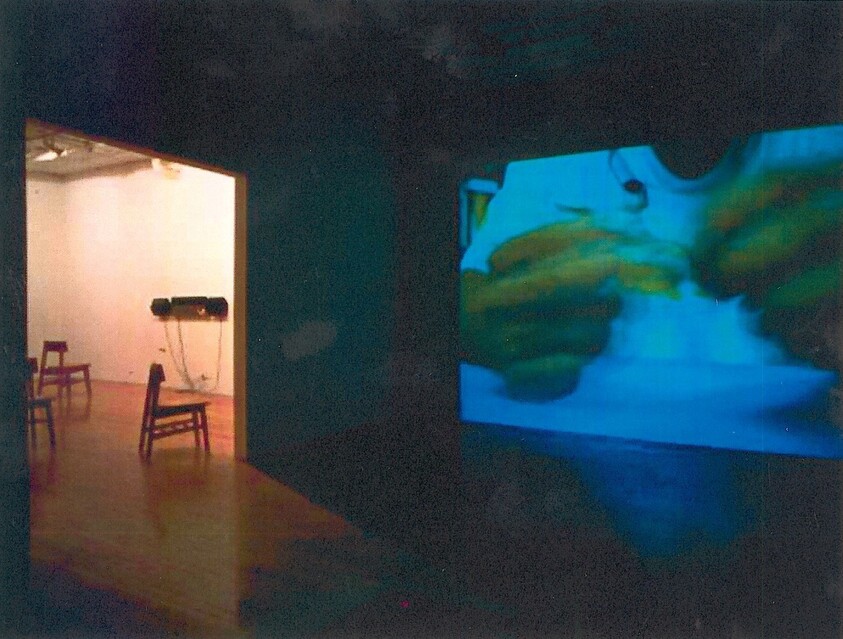 Dark gallery room with a blue video projection of hands and empty chairs in adjacent space.