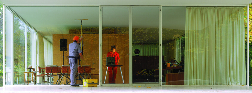 Two workers clean the large glass wall of a modern house, one wiping and one handling equipment.