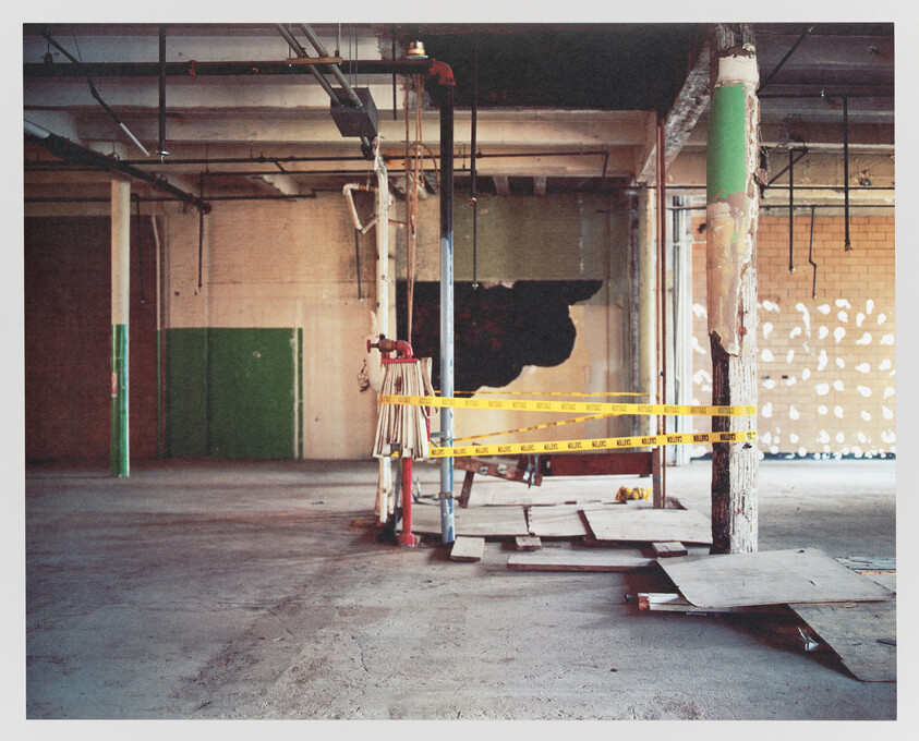 Interior of a dilapidated industrial building with exposed beams, a green door, and caution tape indicating a hazardous area.