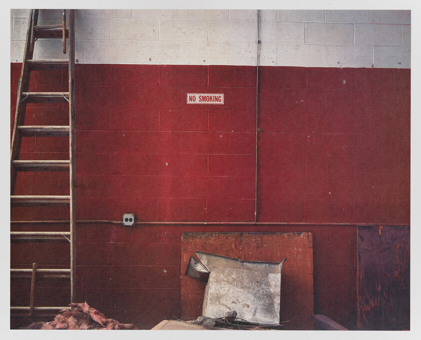 A worn industrial scene with a red wall featuring a "NO SMOKING" sign, a wooden ladder to the left, and various discarded items including a cushion and debris on the floor. The upper part of the wall transitions to gray cinder blocks.