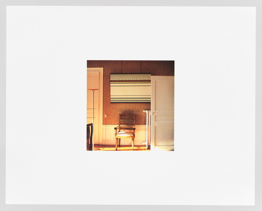 A vintage organ with decorative patterns sits against a wallpapered wall in a warmly lit room, accompanied by a wooden chair with a cushioned seat. The image is centered on a white background, giving the appearance of a photograph within a photograph.