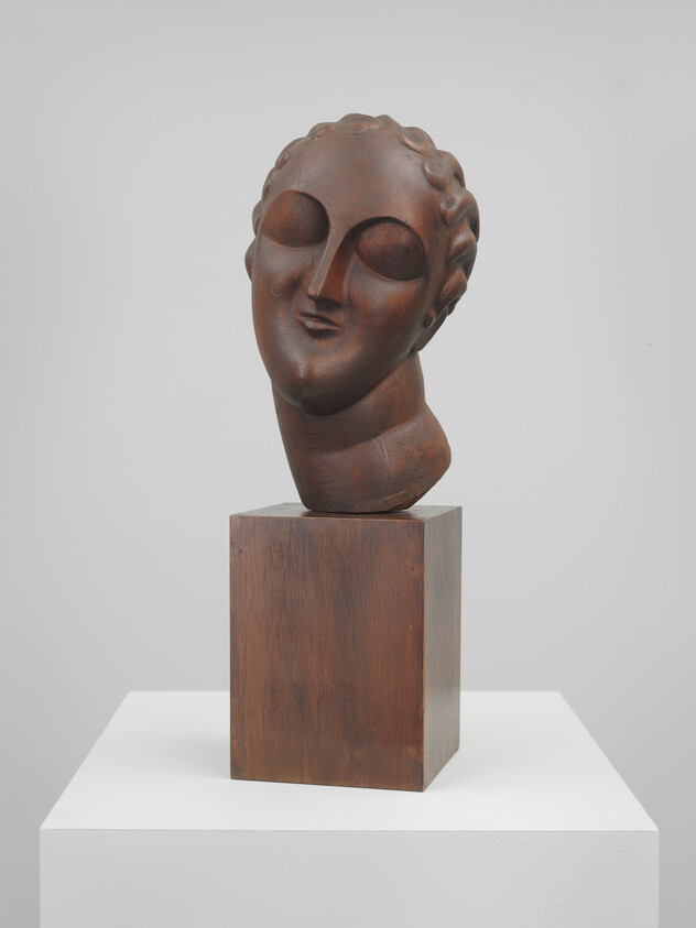 Wooden stylized head sculpture with closed eyes tilted slightly, displayed on a wooden pedestal.