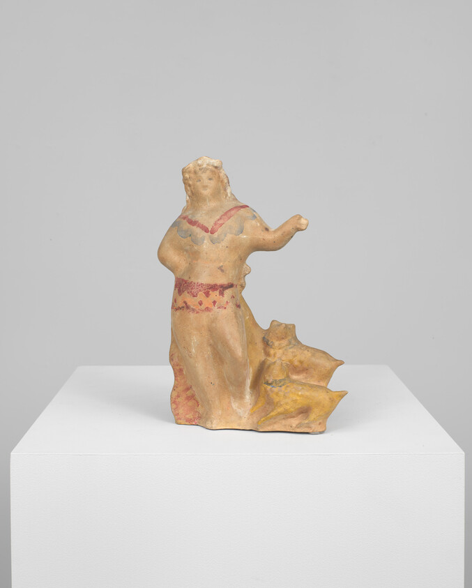 Small painted terracotta figurine of a standing woman with raised arm and flowing drapery on a pedestal.
