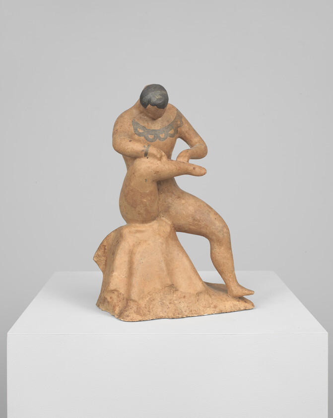 A terracotta figure seated on a rock holding and examining a small animal in its arms.