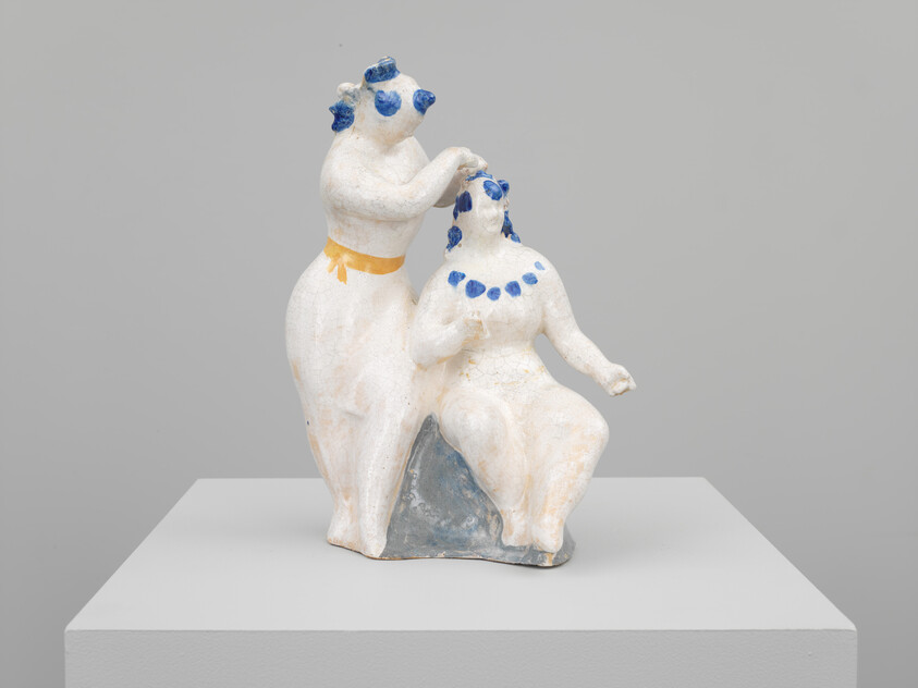 Two white ceramic figures, one standing and grooming a seated figure, with blue dots and a yellow belt.