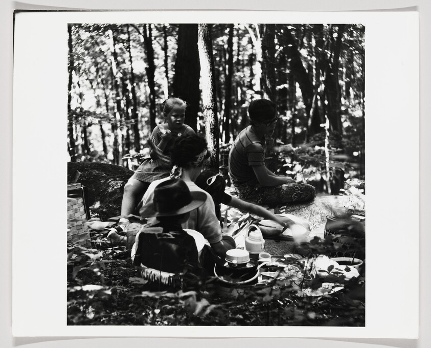 A family sits on the forest floor sharing a picnic with cups, plates, and a thermos.