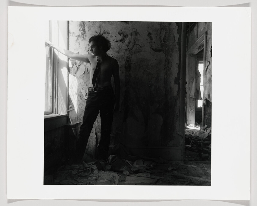 A shirtless young man stands in a dilapidated room looking out a broken window.