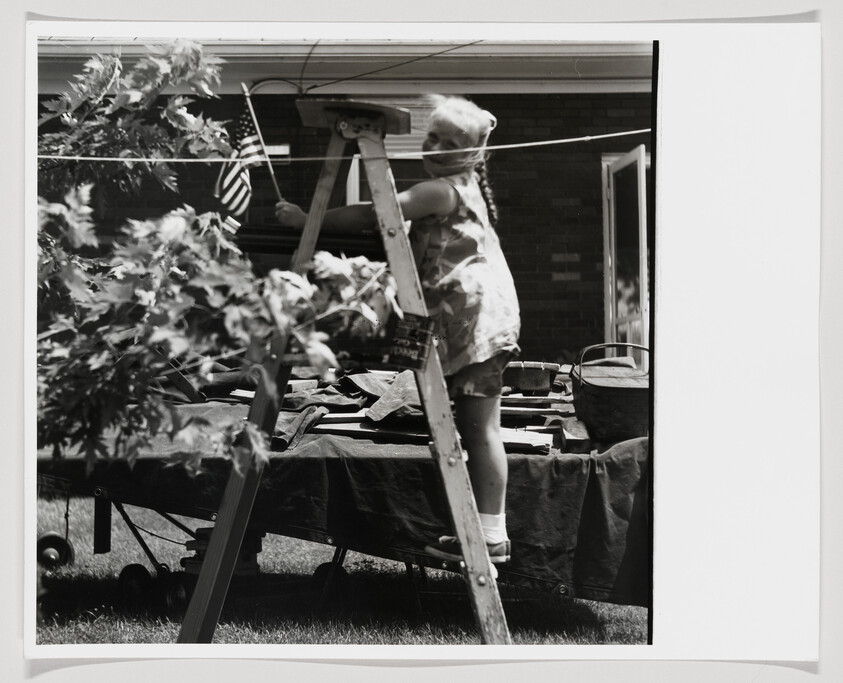 Young girl stands on a ladder holding a small American flag beside a covered table outdoors.