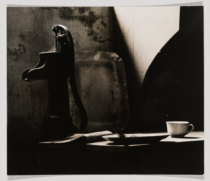 A high-contrast black and white photograph featuring a vintage iron press with its shadow cast on a wall, a blurred object in the background, and a small cup on a saucer in the foreground, all bathed in dramatic lighting that creates sharp shadows.