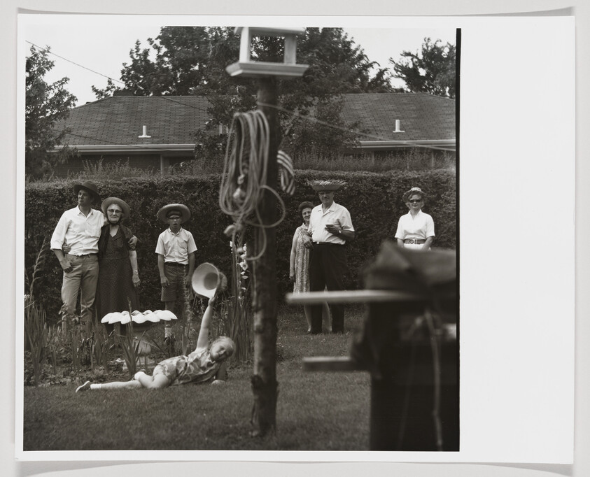 A child lies on the lawn reaching up with a hat while family watches from the garden.