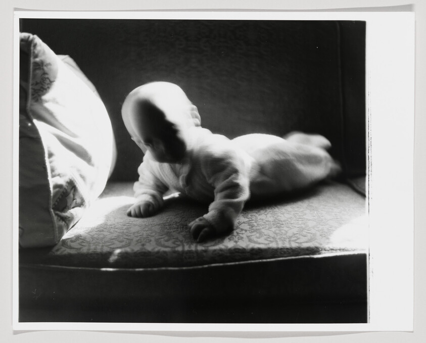 A baby lies on a patterned cushion, propped on hands and bathed in strong side light.