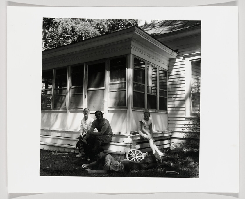 Four people relax on and near a house porch, two sitting on the steps and one lying on the grass.