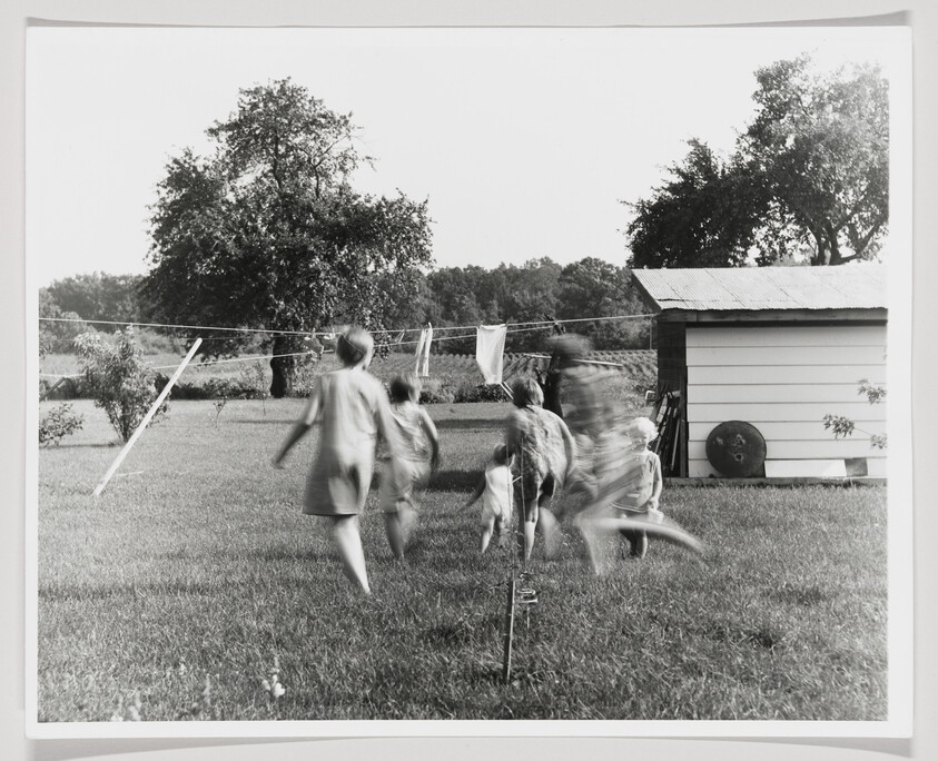 Several children run and play across a backyard lawn past a clothesline and shed.