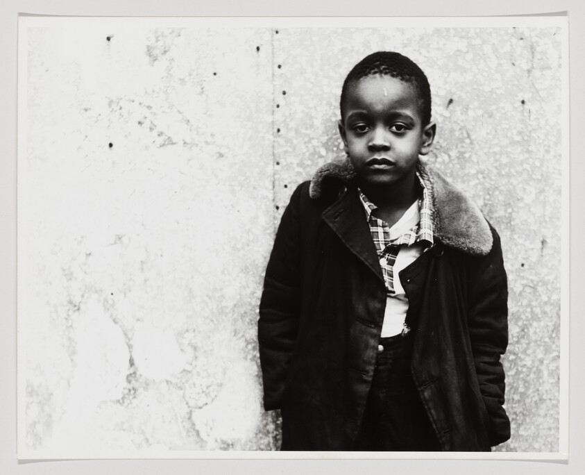 Young boy stands against a textured wall wearing a coat and looking calmly at the camera.