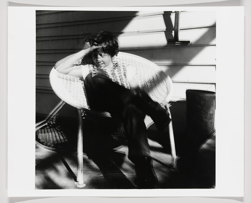 A person sits in a wicker chair on a porch, shading their eyes from bright sunlight.