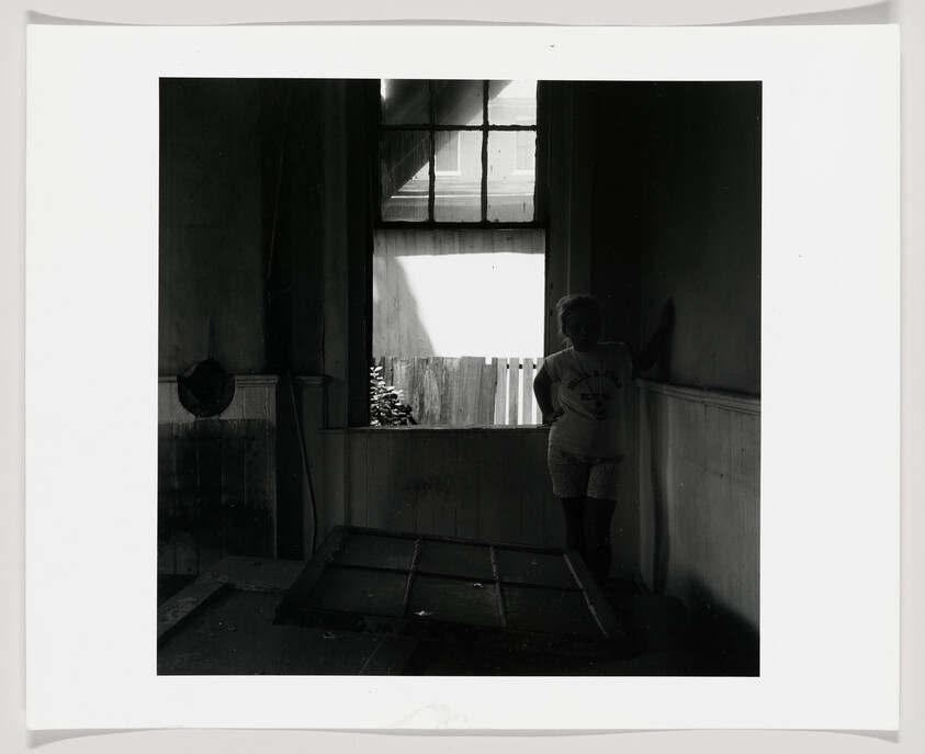 A person stands in a dark room leaning against the wall near a bright window and broken door.