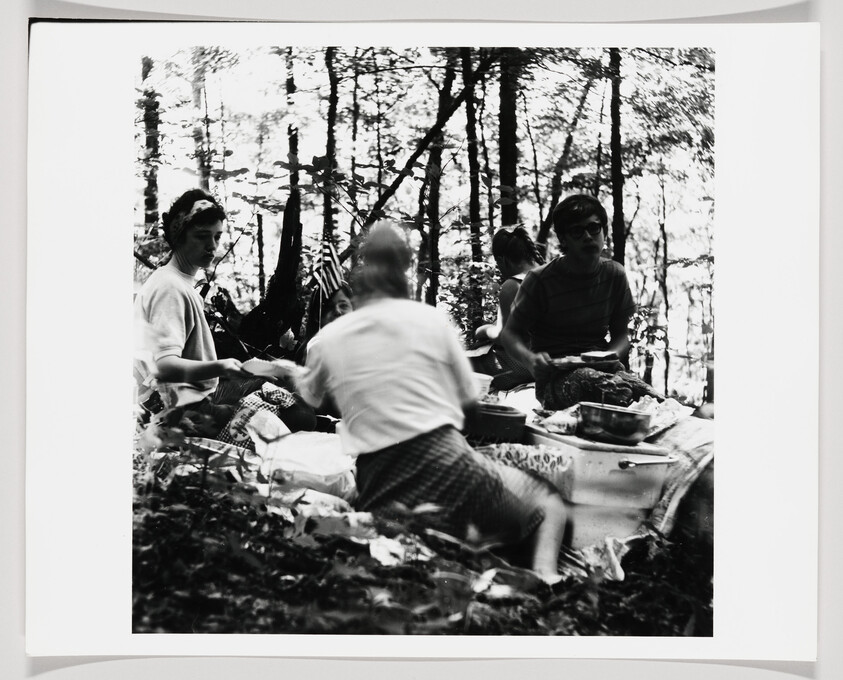 Group of people sit on a blanket in the woods, serving and eating food together.