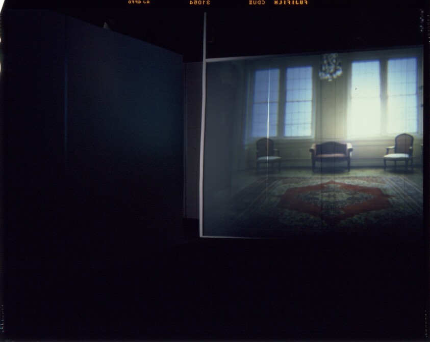 A dimly lit room with a projected image of a bright sitting area, showing chairs, windows, and a rug.
