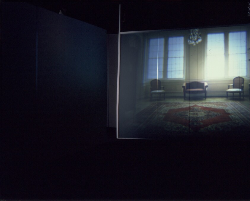 A dimly lit gallery projection showing a room with three chairs and a patterned rug.