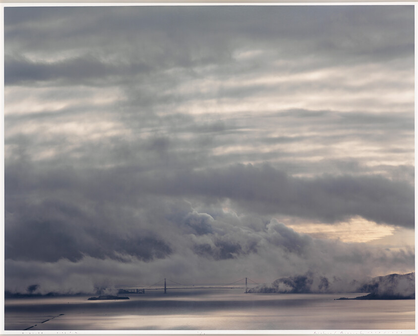 Golden Gate Bridge partially hidden by low fog under a wide, cloudy sky over calm water.